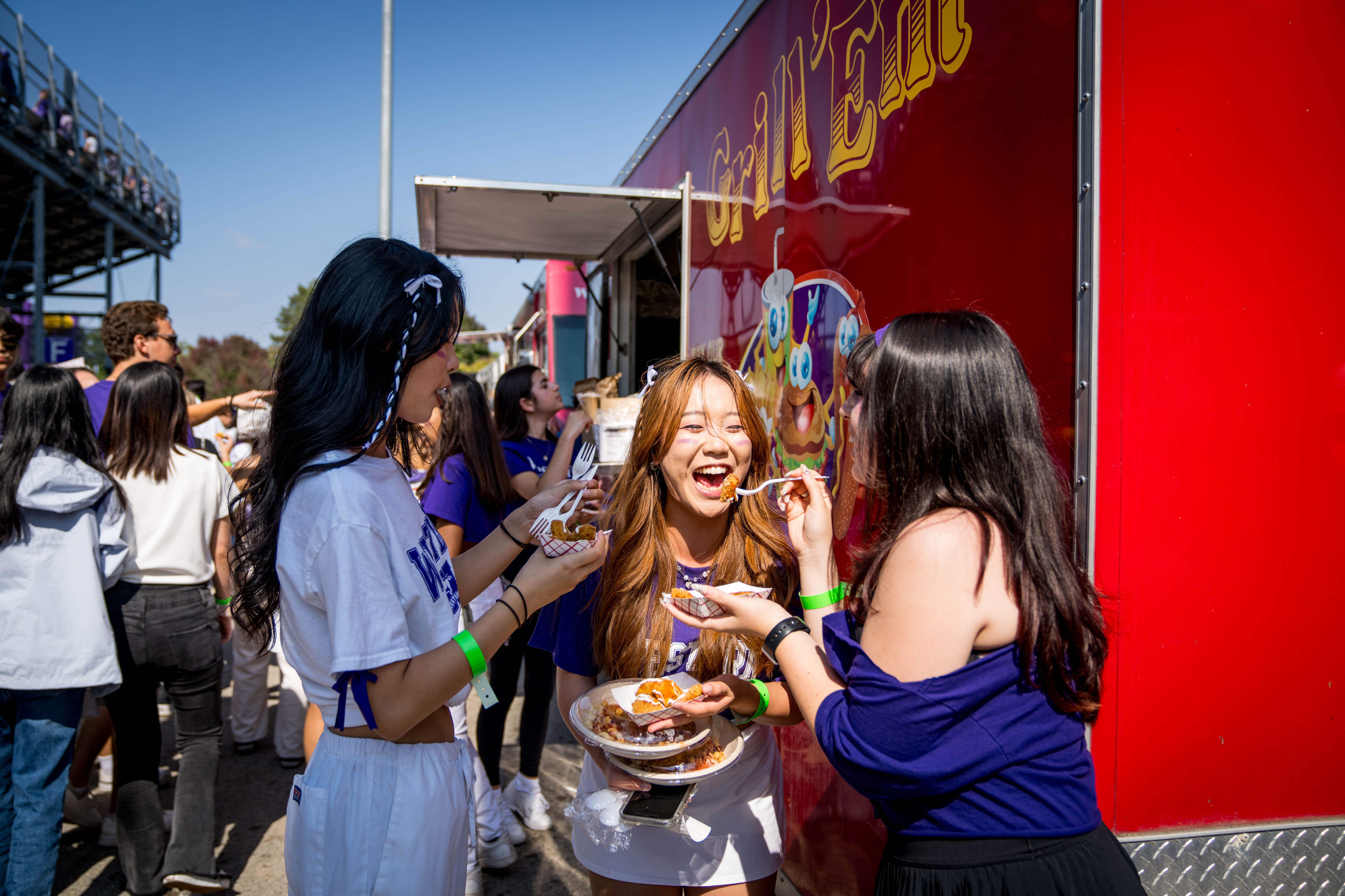 Group of three people eating food outside a food truck while others line up.