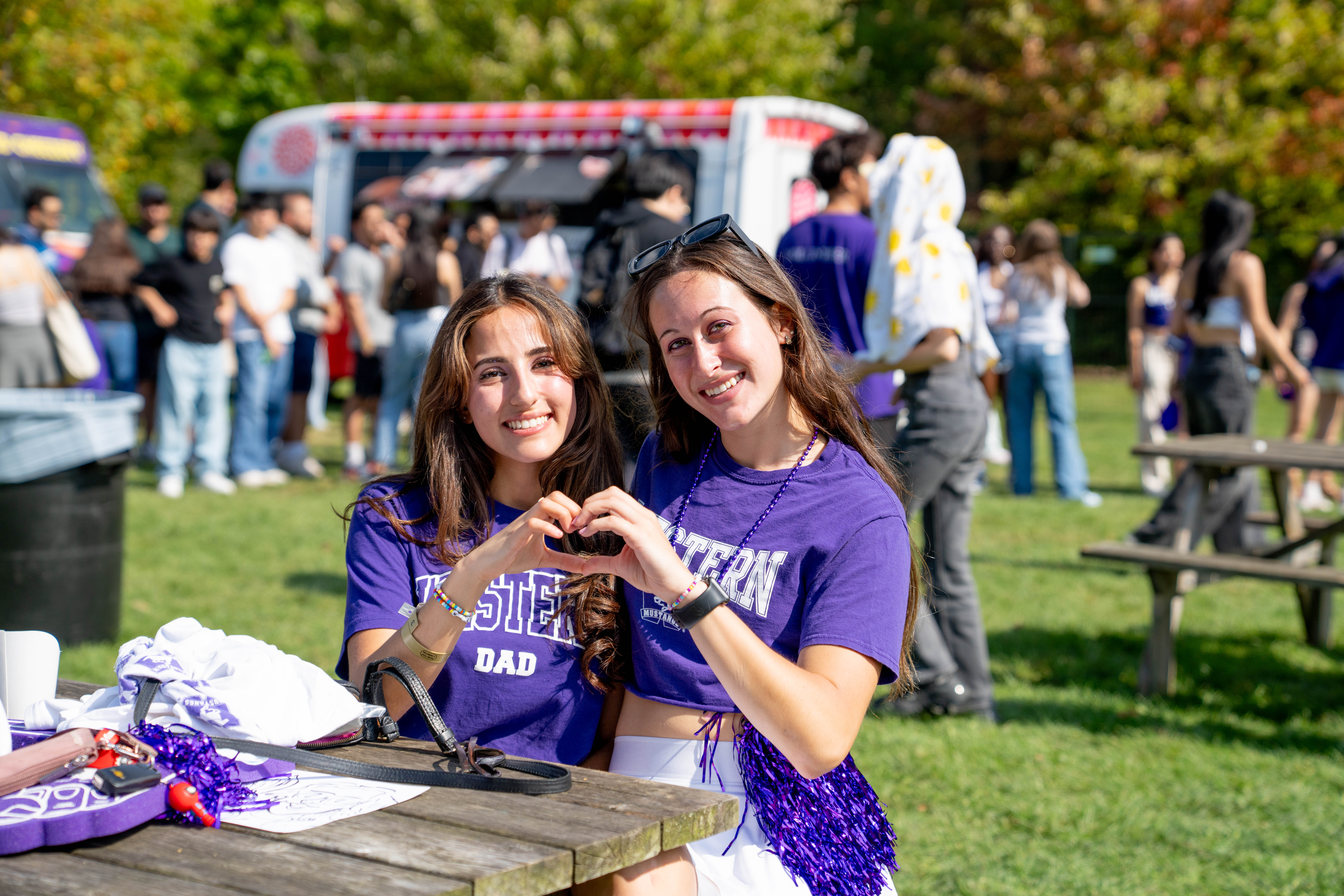 Two People sitting at a bench making a heart sign with their hands with people lined up at food trucks in the background