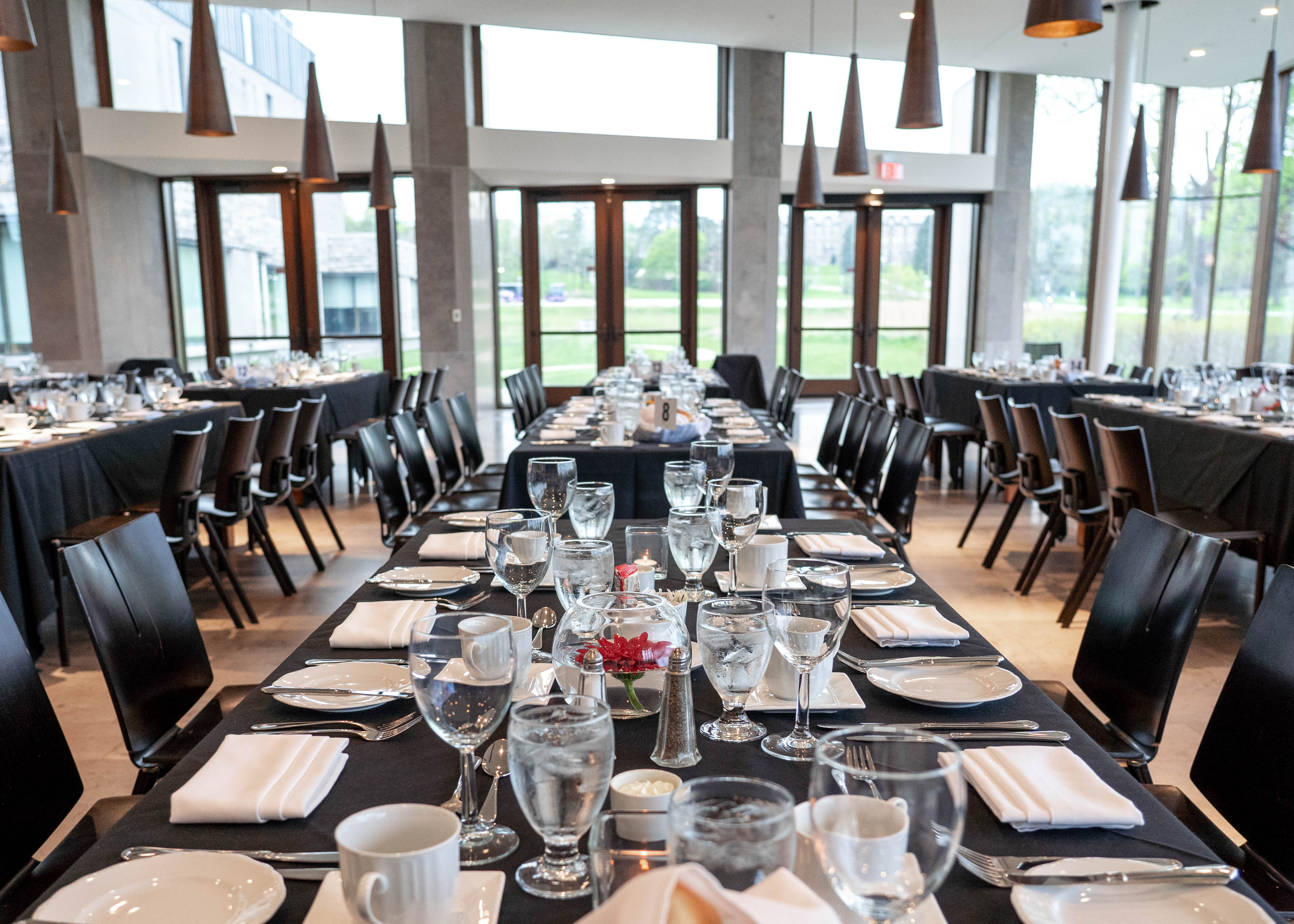 Long banquet tables set with black tablecloths, white plates, folded napkins, and glassware inside a modern event space with floor-to-ceiling windows and pendant lighting.