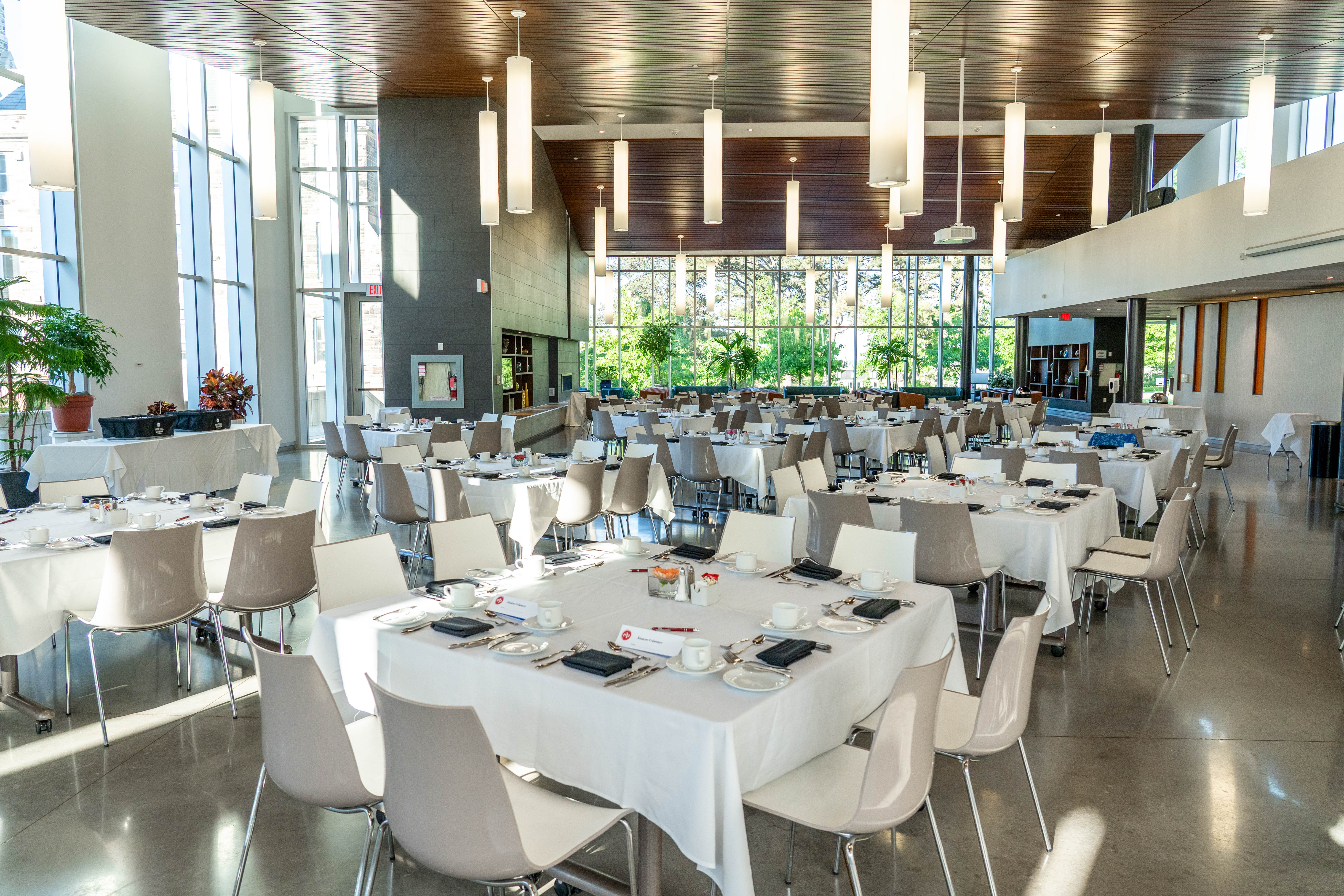 Bright, modern atrium with floor-to-ceiling windows, polished concrete floors, and round tables set for a banquet. Natural light fills the space, which is arranged for a formal event with white tablecloths and neatly placed chairs.